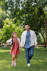 Fototapeta premium Smiling asian parent and child with soccer ball walking in park.