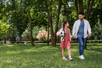 Obraz premium Smiling asian girl holding football and hand of father in park.