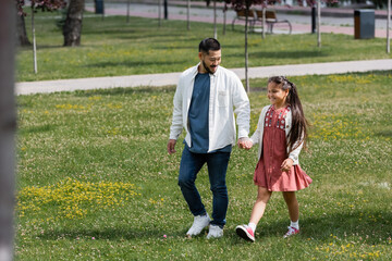 Fototapeta premium Positive asian parent and kid holding hands while walking in park.