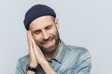 Positive bearded male pretends sleeping, has pleasant dreams, rests on hands, smiles joyfully, wears black stylish hat, isolated over white background. People, good rest and emotions concept.