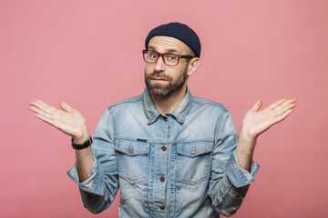Indoor shot of hesitant bearded man shrugs shoulders, looks uncertain, doubts when makes decision,...