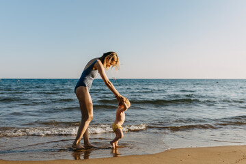 mother helping baby to make first steps. Woman and little child walking on a seashore
