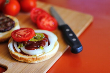 The hamburger is being prepared and served with sliced tomatoes, onions, pickles. on a wooden cutting board in the kitchen