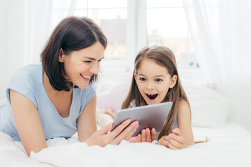 Beautiful mother and daughter watch something funny on tablet computer, connected to wireless internet, spend free time in bedroom, have surprised happy expression. People and leisure concept