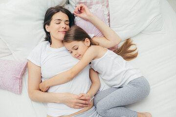 Obraz na płótnie Canvas Family and bed time concept. Pleased sleepy mother and her adorable daughter embrace in bed and have pleasant dreams, lie on white bedclothes, have good relationships, take care of each other