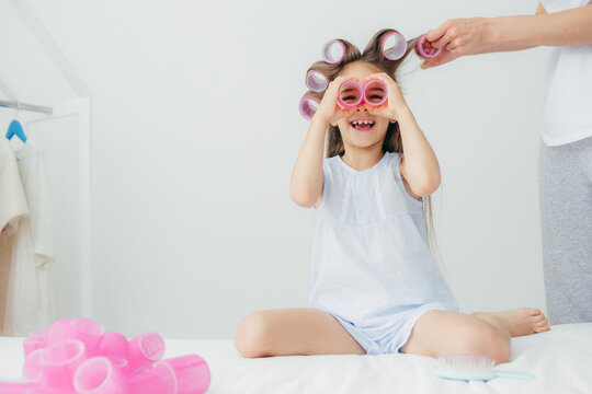Happy cute female child has fun, keeps two hair curlers near eyes, wears nightwear, her mother winds curlers on her long hair, pose in white room. Children, beauty, happiness and joy concept