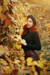Cute red-haired woman covers eye with autumn leaf. Beautiful girl in fall park on walk. Female with black beret, in knitted sweater and leather skirt. Romantic model smiles and looking at camera.