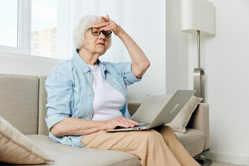 a sad elderly woman is sitting on a sofa in a bright apartment and using a laptop is looking for information on the Internet, holding her hand near her head in frustration, looking with regret