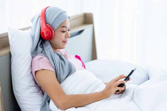 Happy a asian women disease mammary cancer patient with pink ribbon wearing headscarf in headphones is listening to music with smartphone After treatment to chemotherapy sit on bed In the bedroom
