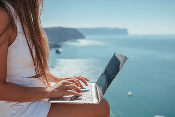 Digital nomad, woman in the hat, a business woman with a laptop sits on the rocks by the sea during sunset, makes a business transaction online from a distance. Freelance, remote work on vacation.