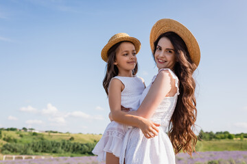 brunette woman in straw hat smiling at camera while holding daughter in countryside.