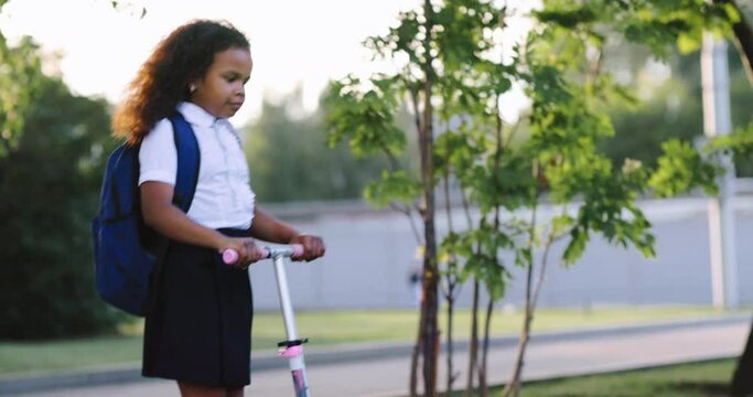 African american girl teenager in school uniform and a school backpack rides a scooter on a summer day in the park to school