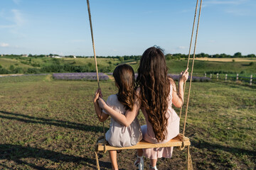 back view of long haired woman and girl riding swing in countryside.