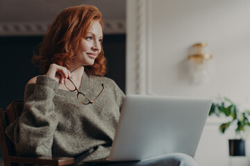 Experienced redhead female entrepreneur makes plans, looks aside with dreamy expression, uses modern technology, holds spectacles, wears casual jumper, enjoys distance education and e learning
