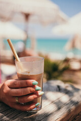 Woman hand with turquoise manicure hold ice coffee latte in tall glass with straw by the sea in beach bar