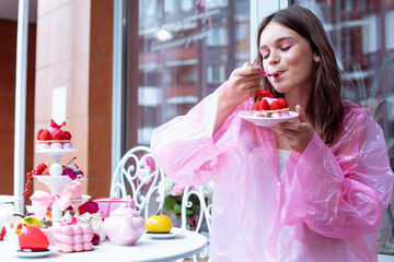 Pretty teenage girl in pink raincoat joying, tasting sweet berry dessert cakes from the table in...