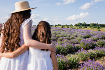 back view of brunette mom and girl in straw hats embracing in lavender meadow.