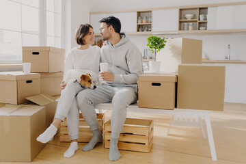 Lovely spouses sit near carton big boxes, drink coffee and talk about future plans, relocate in new...