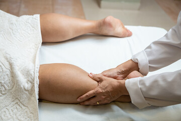 Uma terapeuta vestida de branco, fazendo massagem na perna de paciente deitado em maca. - obrazy, fototapety, plakaty