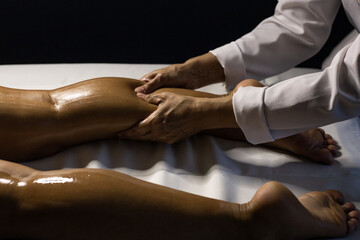 Uma terapeuta vestido de branco, fazendo massagem na perna de paciente deitado em maca, com o uso de óleo terapêutico. - obrazy, fototapety, plakaty