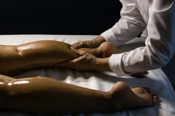 Uma terapeuta vestido de branco, fazendo massagem na perna de paciente deitado em maca, com o uso de óleo terapêutico. - obrazy, fototapety, plakaty
