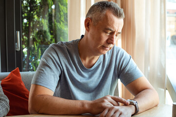 Portrait of thoughtful elderly man sitting at table on sofa near window, looking at watch, waiting...