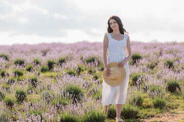 happy woman in summer dress holding straw hat while standing in lavender field.