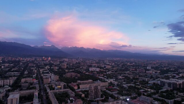 Pink-blue large clouds over the city of Almaty and the mountains. Top view from a drone. The houses are illuminated with pink light from the sunset. In the distance there are green hills and TV tower