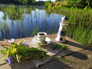Still life with coffee breakfast, lighthouse toy, forest flowers on rustic wooden pier on riverside in sunny summer morning