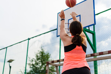 Woman in a bright tank top shooting a basketball at an outdoor court, with focus on determined posture and the basketball aimed towards the hoop, set against a clear sky.