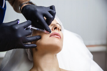 The doctor cosmetologist in black gloves makes Lip augmentation procedure of a european woman in a beauty salon. 