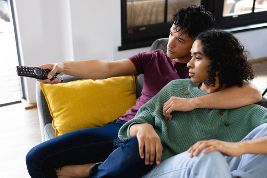 Happy biracial lgbt male couple embracing on sofa in living room, watching tv