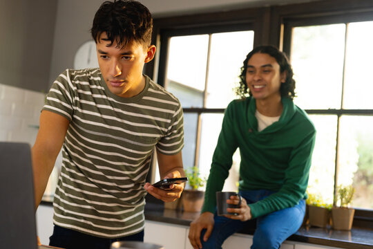 Happy biracial lgbt male couple using laptop and smartphone in kitchen together
