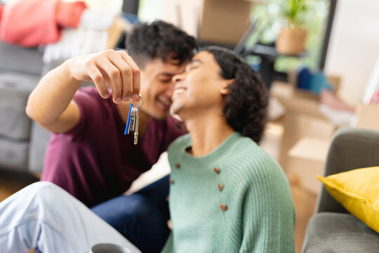 Happy biracial lgbt male couple moving house, sitting on floor in living room holding house keys