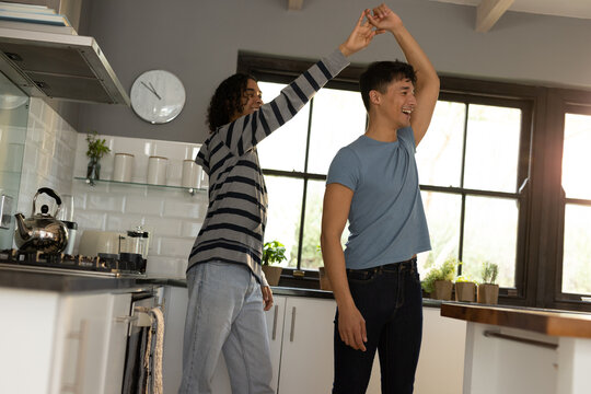 Happy biracial lgbt male couple in kitchen, dancing and smiling