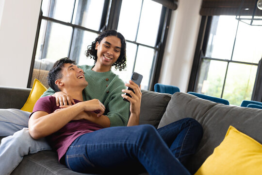 Happy biracial lgbt male couple sitting on sofa in living room, embracing and using smartphone