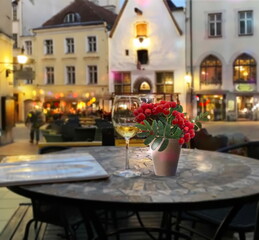 Fototapeta na wymiar Evening street cafe glass of wine and red rowan ash berry branch on table , people walk evenight blurred light vestive decoration in medieval city Tallinn old town