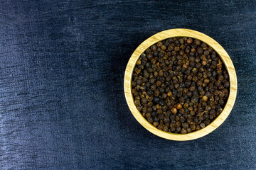 Black pepper in wooden bowl on black wooden background, top view. 