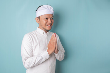 Smiling young Balinese man wearing udeng or traditional headband and white shirt gesturing greeting or namaste isolated over blue background - obrazy, fototapety, plakaty