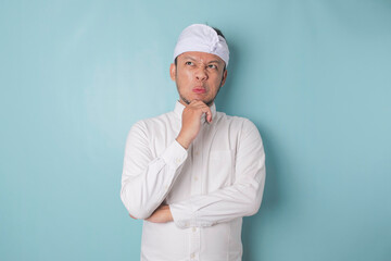 Portrait of a thoughtful young Balinese man wearing udeng or traditional headband and white shirt looking aside while his finger on his chin isolated over blue background