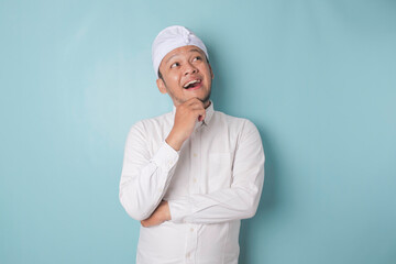 Portrait of a thoughtful young Balinese man wearing udeng or traditional headband and white shirt looking aside while his finger on his chin isolated over blue background - obrazy, fototapety, plakaty