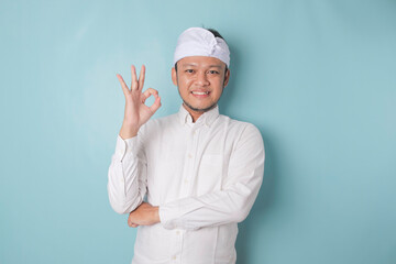 Excited Balinese man wearing udeng or traditional headband and white shirt giving an OK hand gesture isolated by a blue background