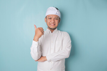 Excited Balinese man wearing udeng or traditional headband and white shirt gives thumbs up hand gesture of approval, isolated by blue background