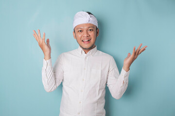 Excited Balinese man wearing udeng or traditional headband and white shirt pointing at the copy space upside him, isolated by blue background