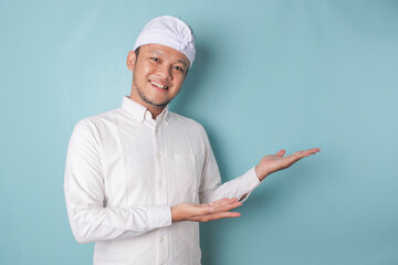 Excited Balinese man wearing udeng or traditional headband and white shirt pointing at the copy space beside him, isolated by blue background