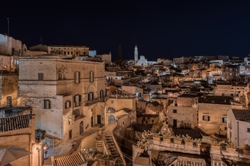 Fototapeta na wymiar Matera by night, photos from the panoramic terrace. Photo from June 2022. You can see the basilica overlooking the town and the characteristic houses carved into the rock. Matera was the European capi