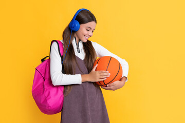 positive child in headphones with school backpack and basketball ball on yellow background