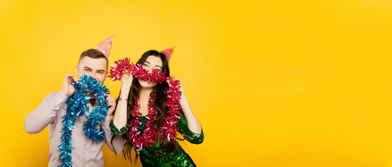Young adult boy and girl 20-25s in party hats pose with New Year's tinsel, make mustaches and look at the camera on a yellow isolated background. Festive banner with copy space