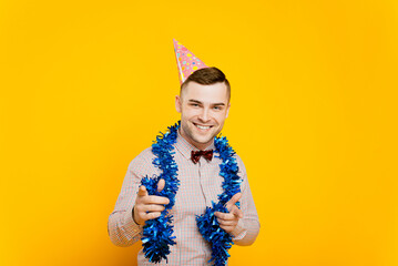 Young adult guy 20-25s wearing a party hat and new year tinsel making a gesture pointing with index...