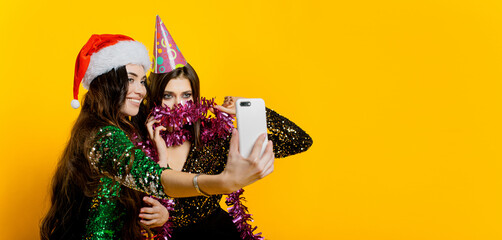 Young adult girls 20-25s in a party hat and santa claus cap and tinsel make a video or take a selfie for social networks on a yellow isolated background. Banner for new year party
